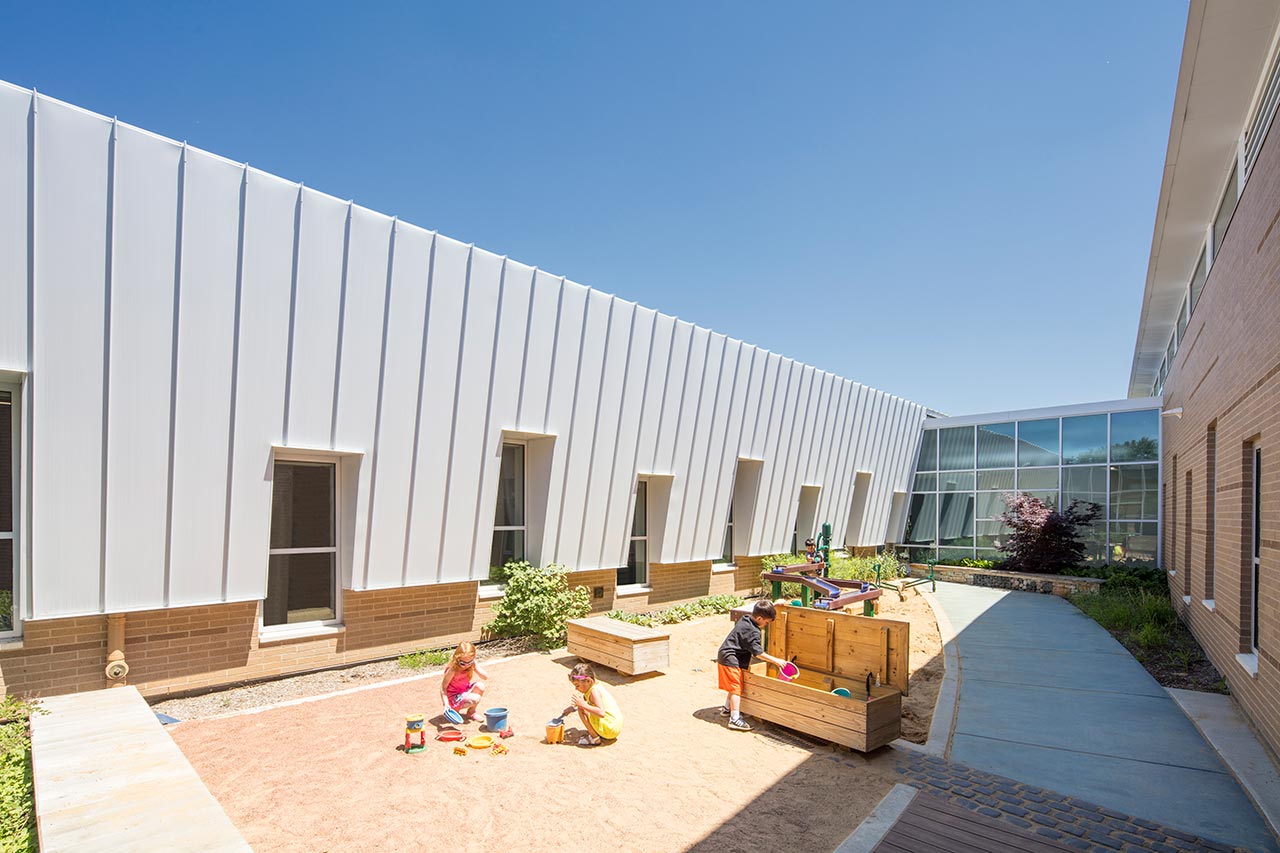 Early learners playing in school courtyard with metal wall and random windows in background