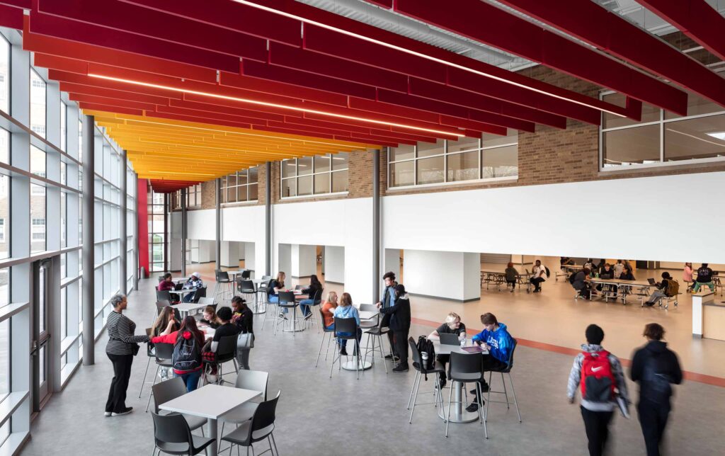 High school students in two-story commons with red and yellow ceiling, glass wall on left, and cafeteria (lower lever) and classrooms (upper level) on right.