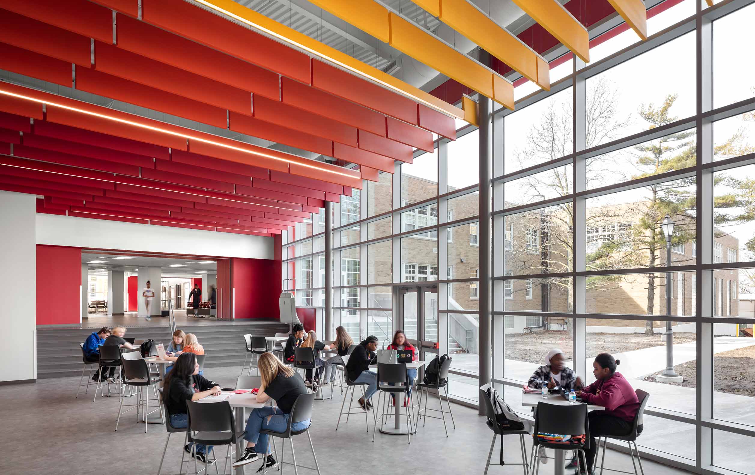 High school students in commons with red and yellow ceiling highlights and glass wall displaying courtyard