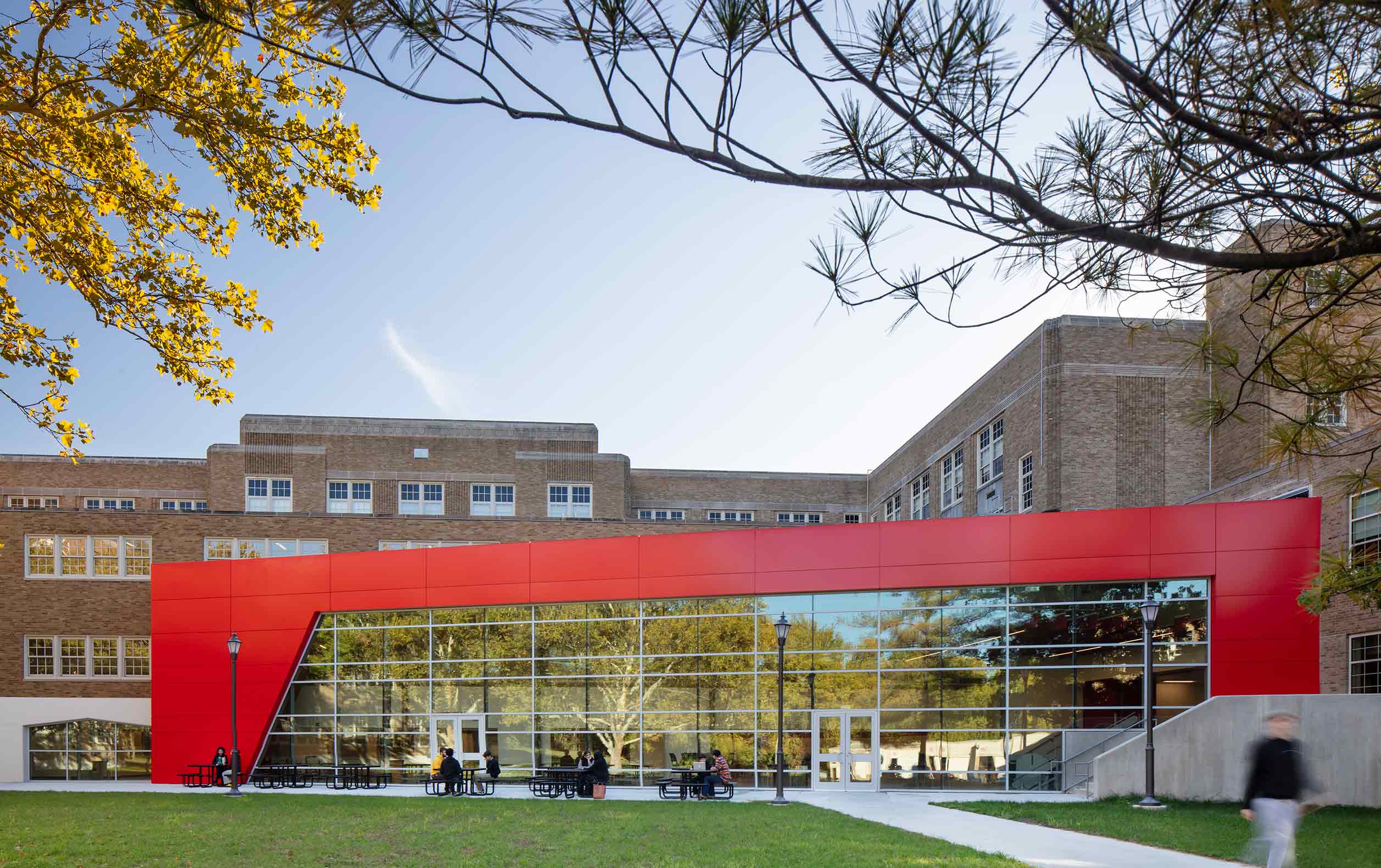 High school students at tables in courtyard. Background is school addition with red border and glass wall reflecting trees.