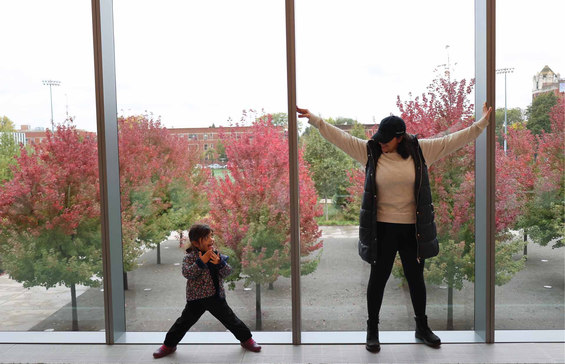 Woman and child pose playfully in front of floor-to-ceiling windows overlooking red autumn trees.