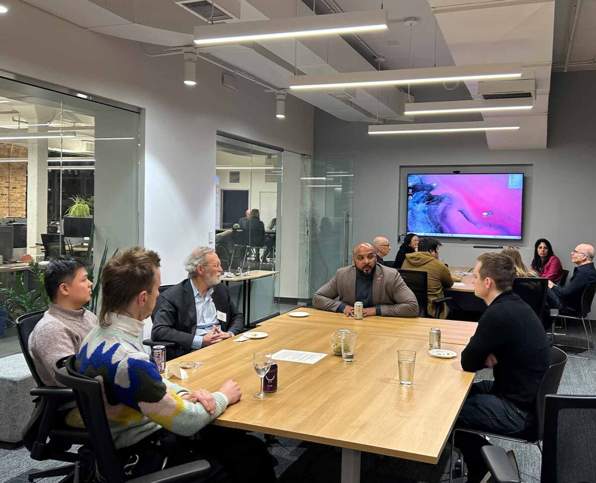 A group of people sit around a conference table in a modern office, engaged in discussion with a screen in the background.