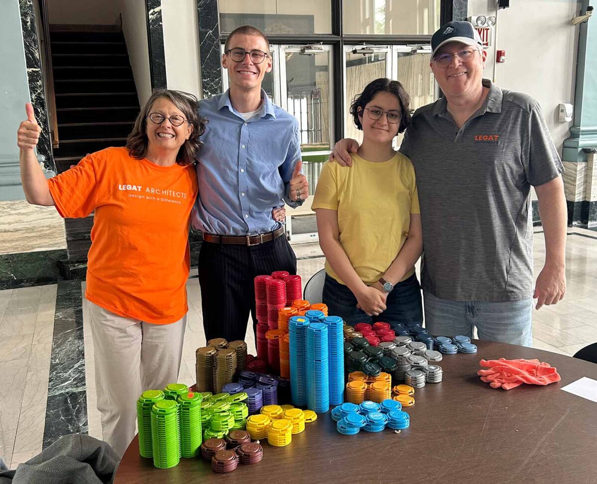 Four people stand behind a table filled with neatly stacked colorful plastic pieces, smiling and giving thumbs up.