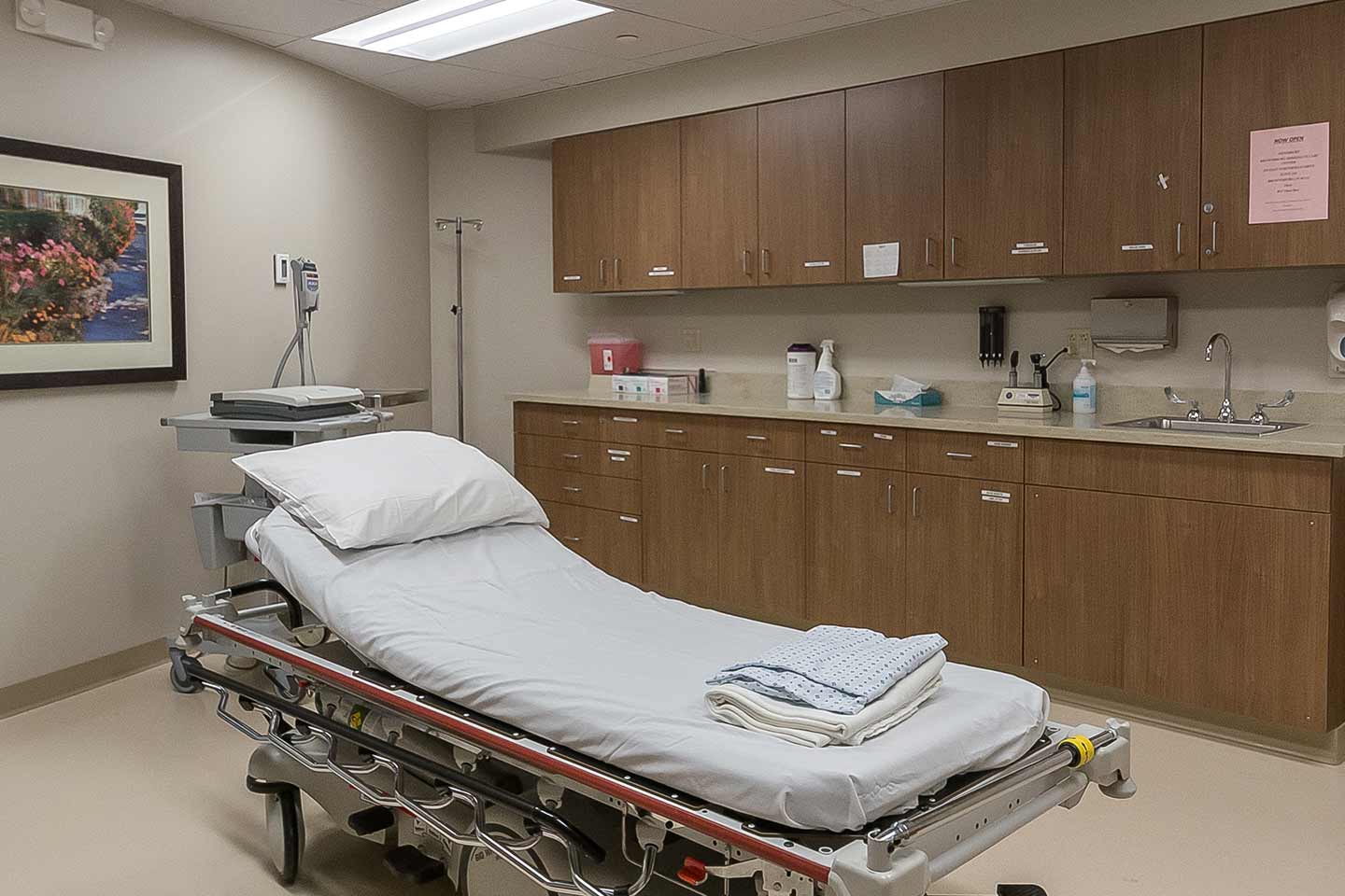 Patient exam room with hospital bed, medical equipment, cabinets, and sink in a modern healthcare facility.