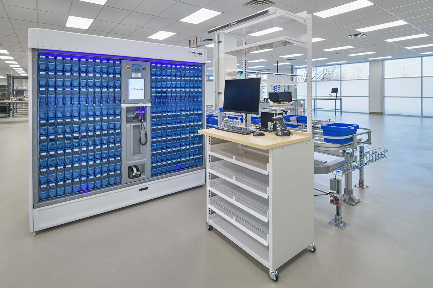 Automated medication storage system with conveyor, workstation, and blue bins in a modern pharmacy facility.