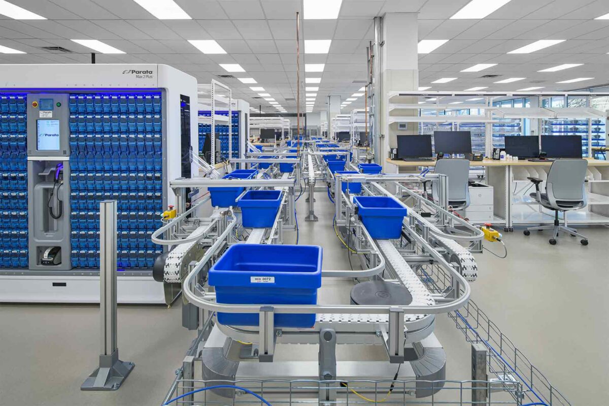 Automated warehouse conveyor system with blue storage bins and workstations in a modern fulfillment center.