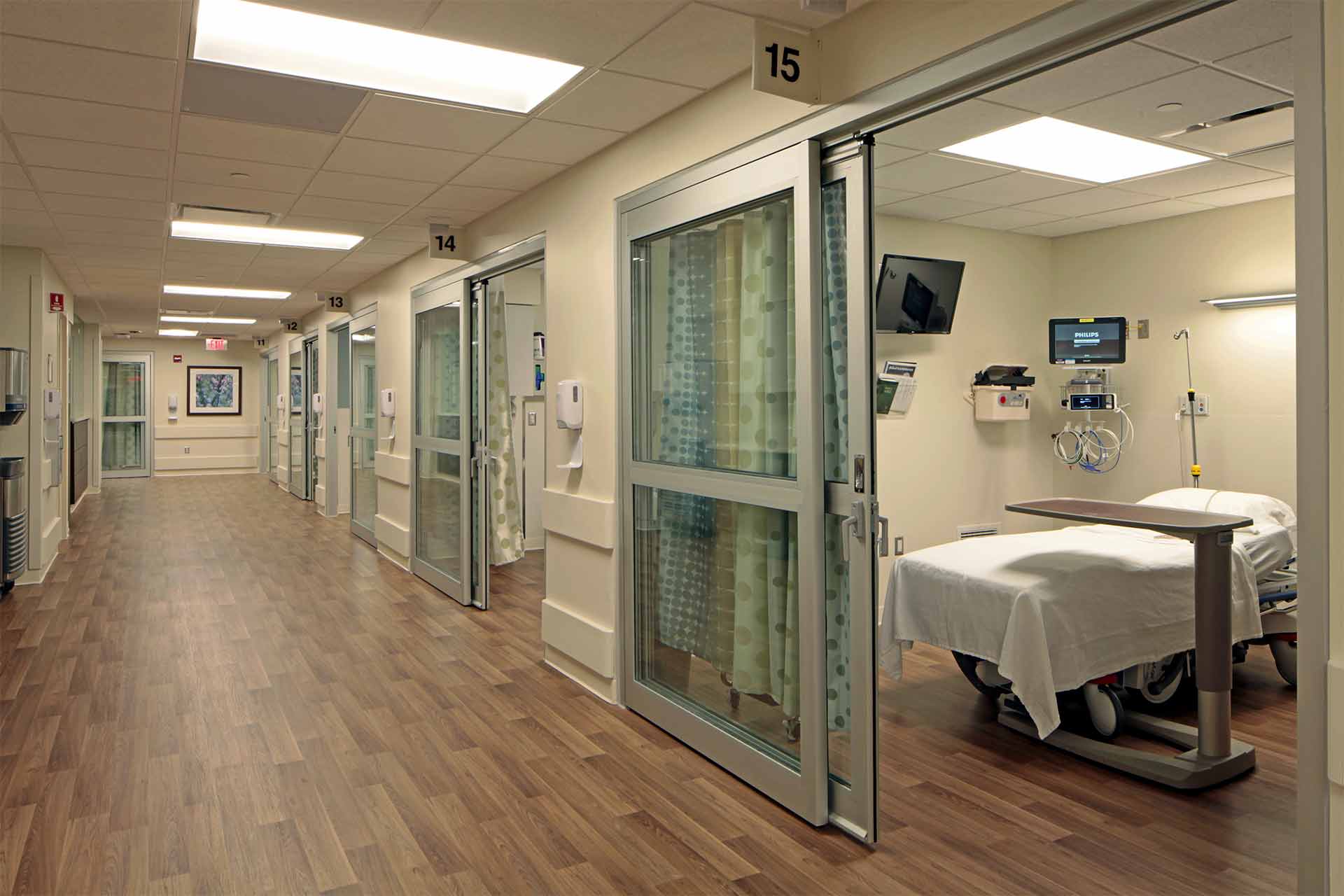 Hospital corridor with private patient rooms, medical equipment, and an open treatment room in a modern care facility.