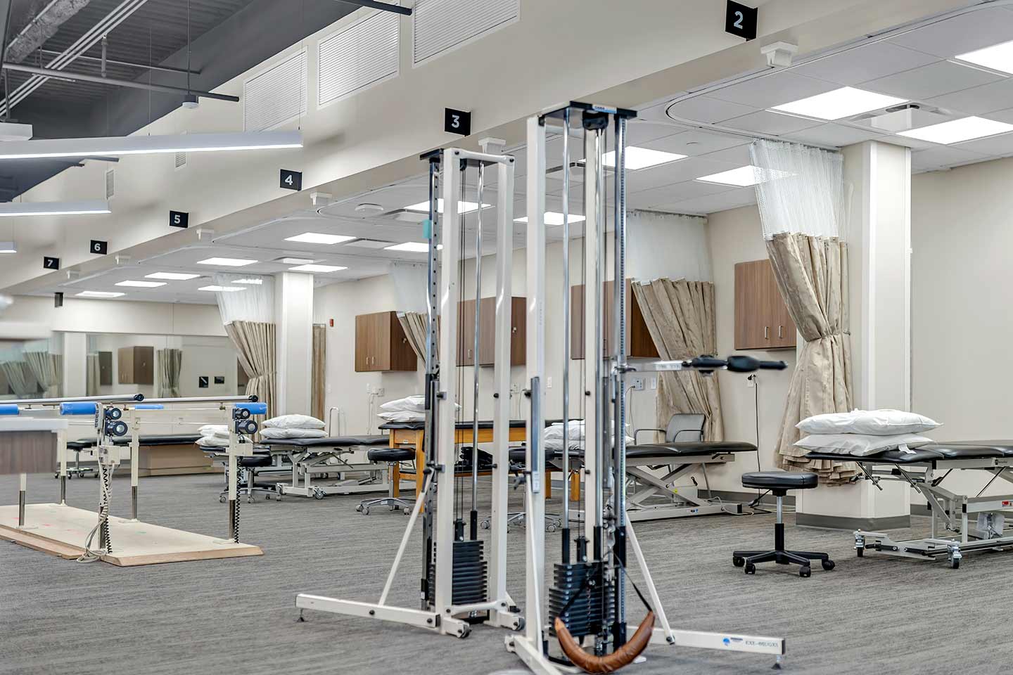 Modern rehabilitation therapy gym with treatment tables, exercise equipment, and curtained patient care stations.