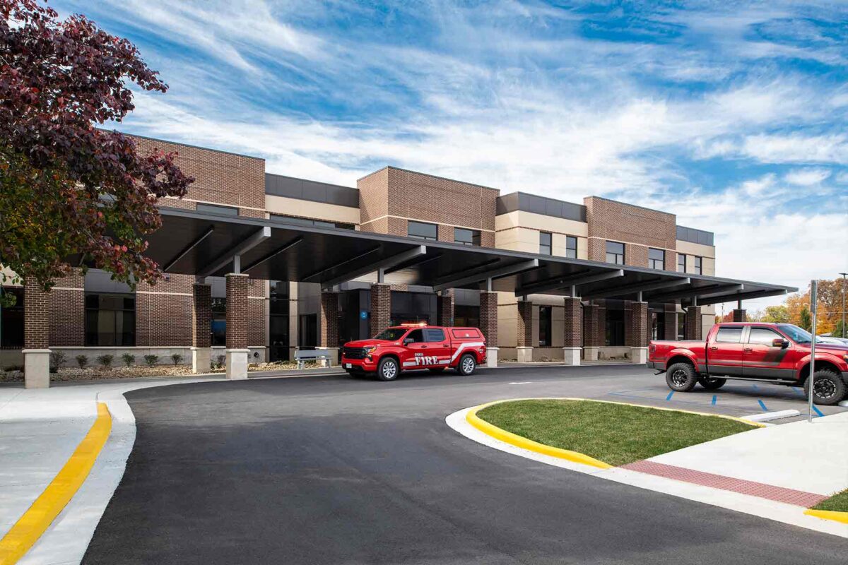 Modern hospital entrance with covered drop-off, fire department vehicle, and clear blue sky.