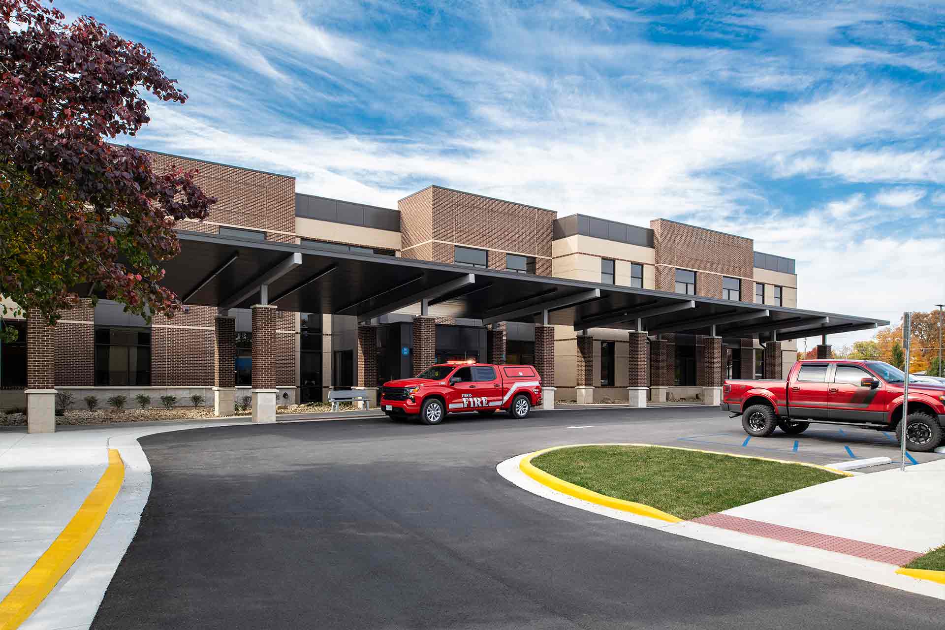 Modern hospital entrance with covered drop-off, fire department vehicle, and clear blue sky.