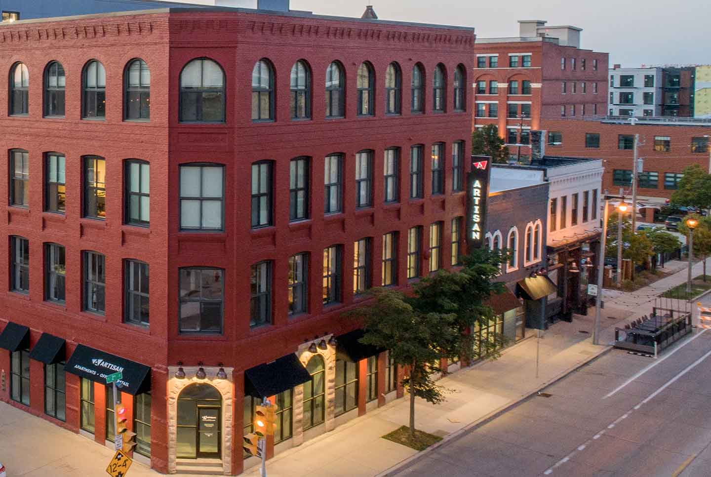 Exterior view of a red brick building called Artisan with arched windows along a city street at dusk.