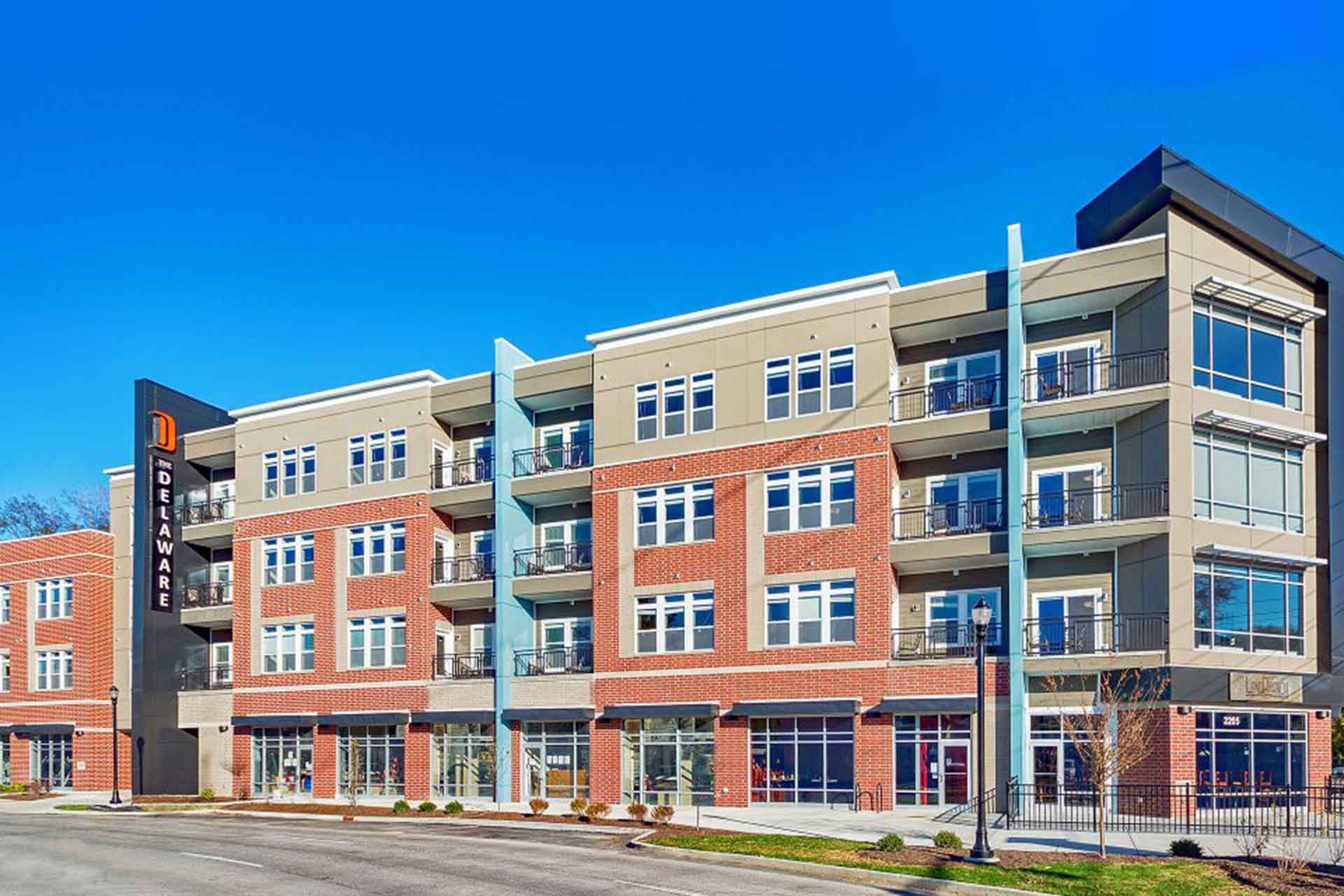 Exterior view of The Delaware apartment building featuring red brick, blue accents, balconies, and retail storefronts.