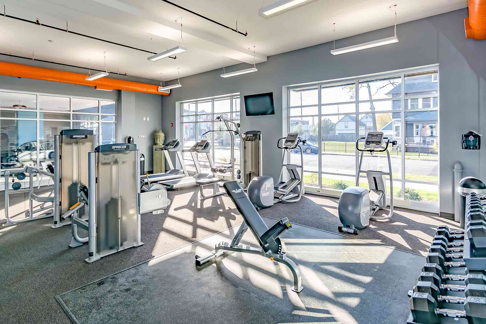 Bright fitness center featuring exercise machines, weight benches, free weights, and large windows with street views.
