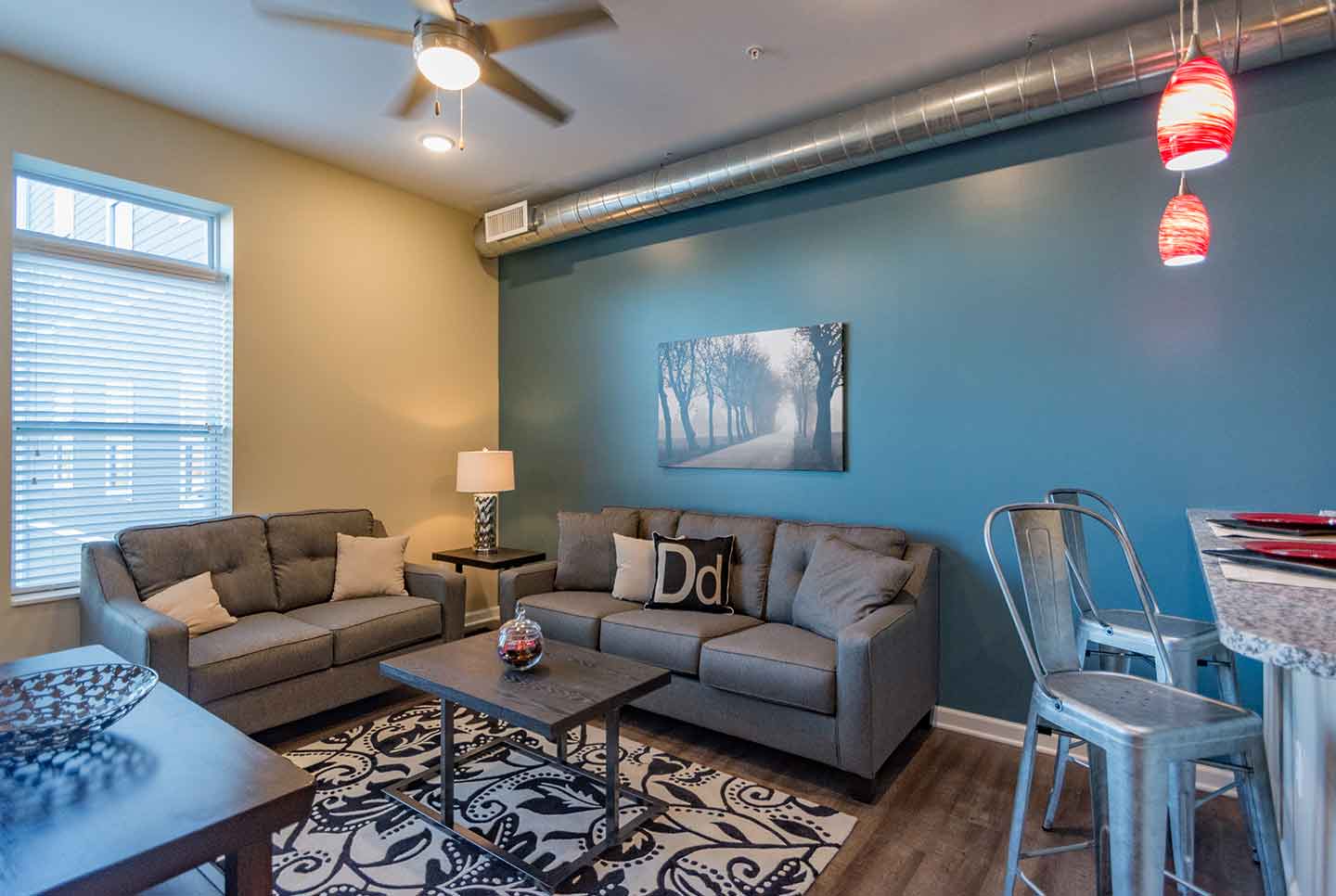 Modern apartment living room with teal accent wall, grey sofas, pattern rug, exposed ductwork, and kitchen island.