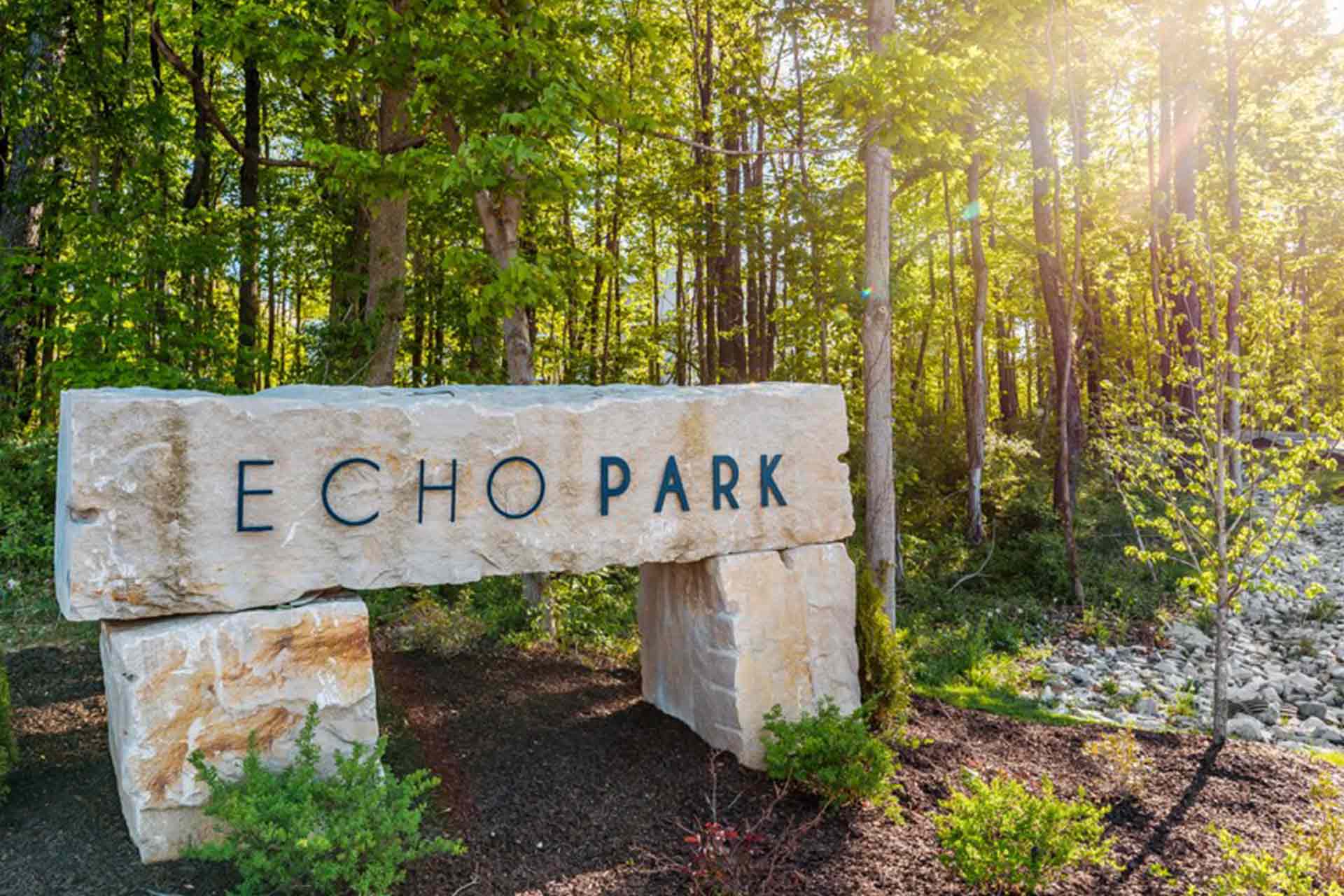 Large stone monument sign reading "Echo Park" set against a lush, sunlit wooded backdrop.