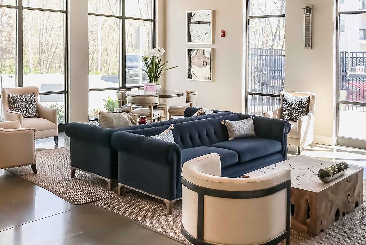 Modern apartment lounge featuring dark blue tufted sofas, cream armchairs, large windows, and contemporary decor.