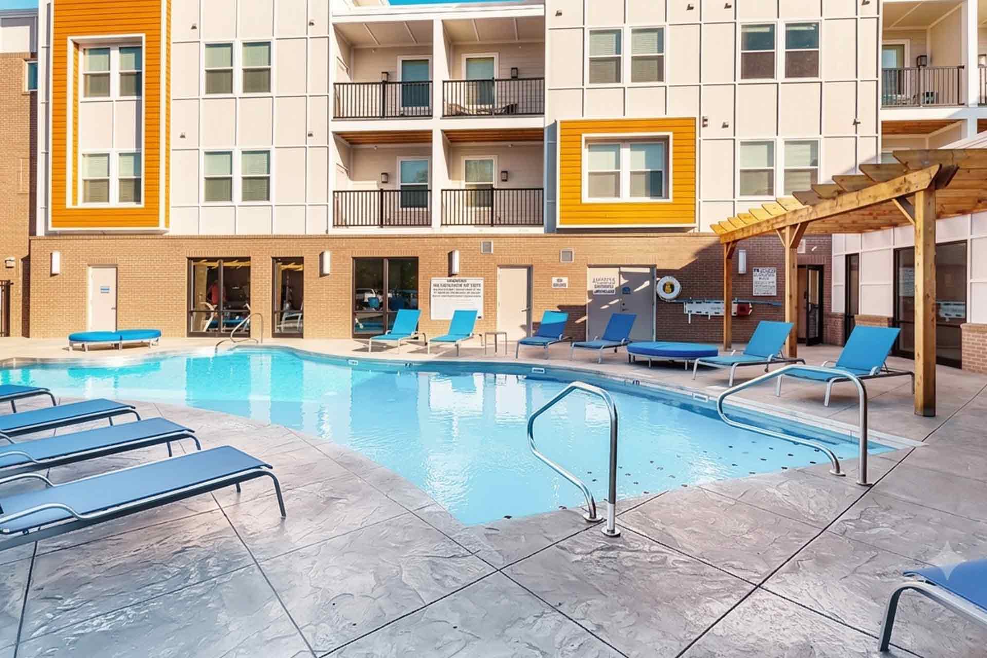 Outdoor swimming pool area with blue lounge chairs and wooden pergola outside a modern apartment building.