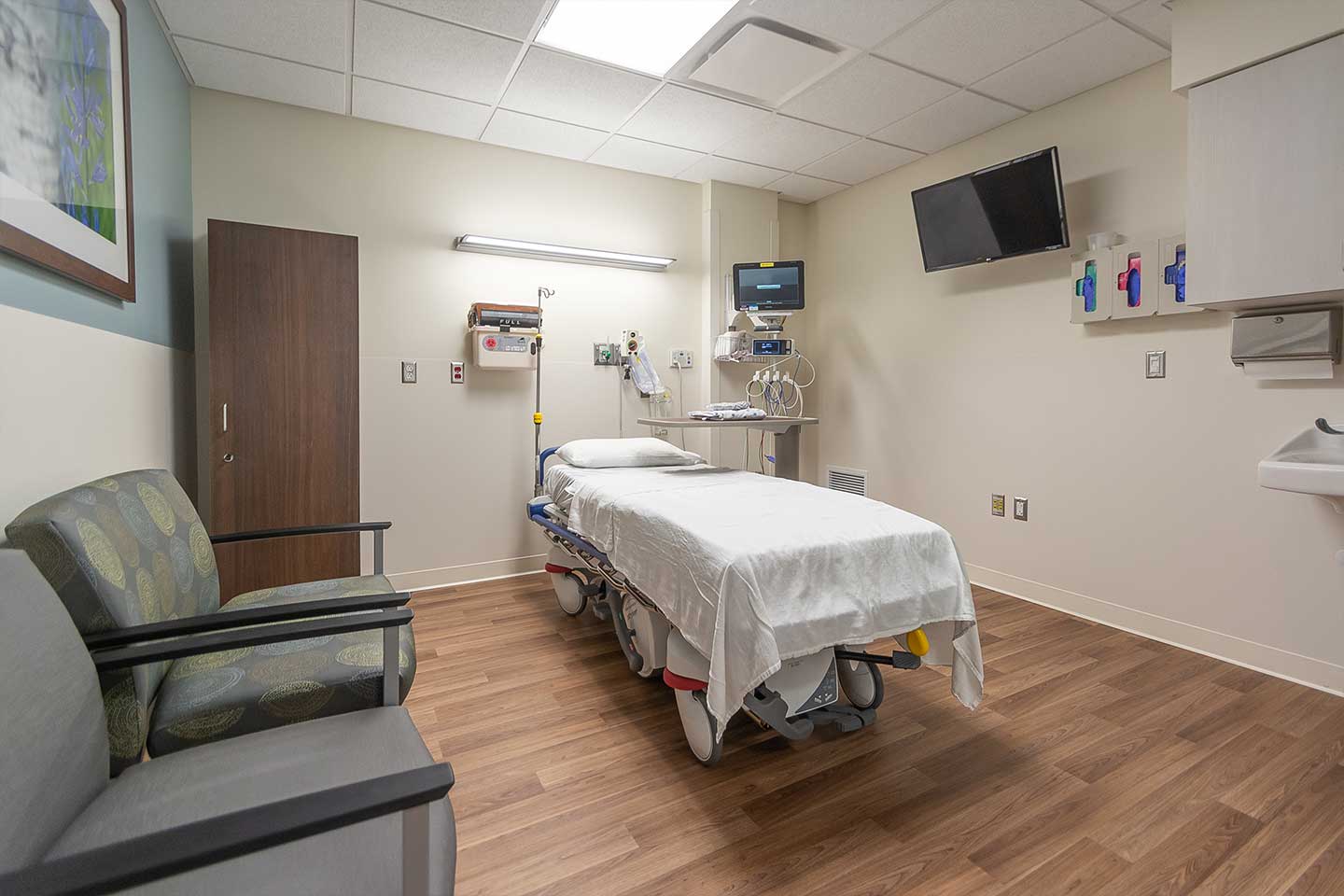 Modern hospital patient room with exam bed, medical monitors, seating, and clinical equipment.