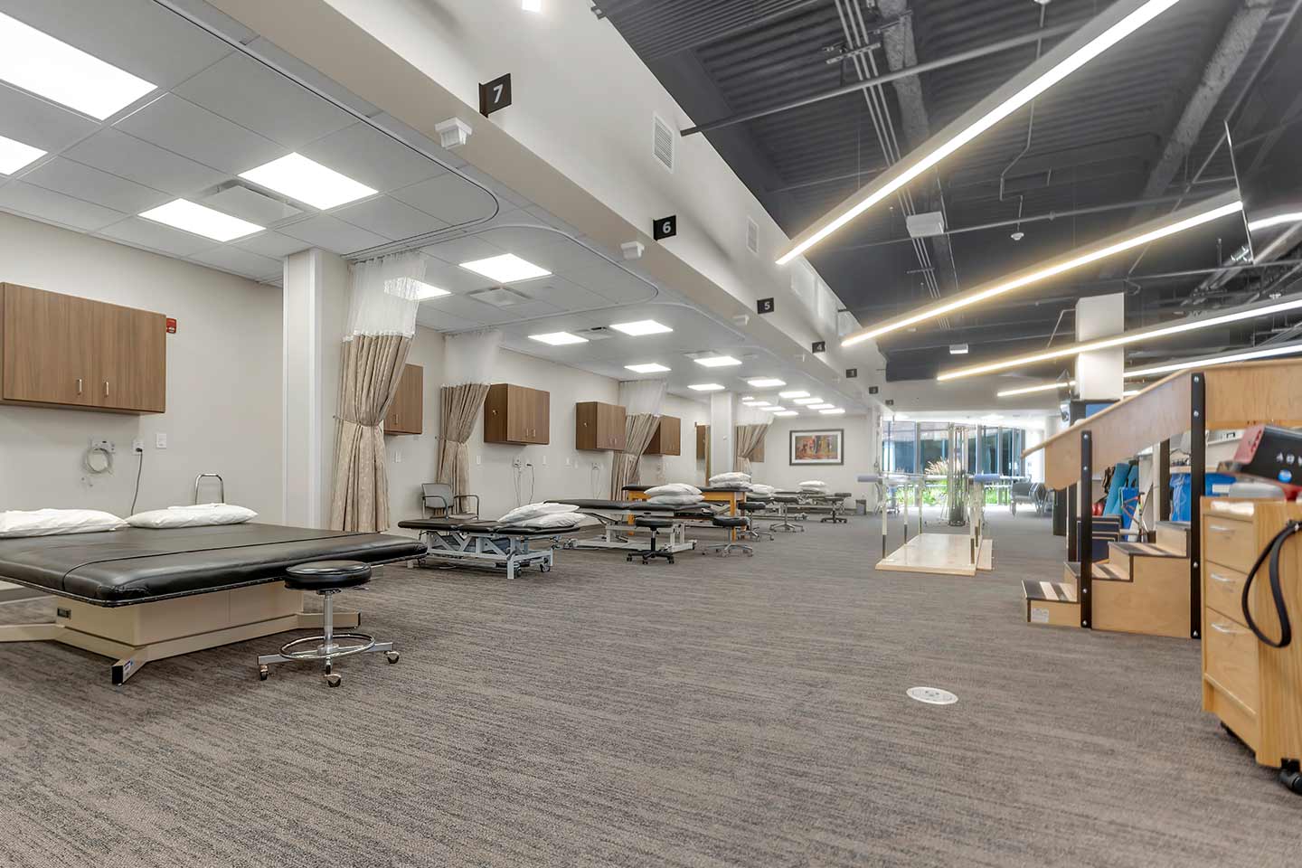 Open rehabilitation therapy gym with treatment tables, exercise equipment, and curtained patient care bays.