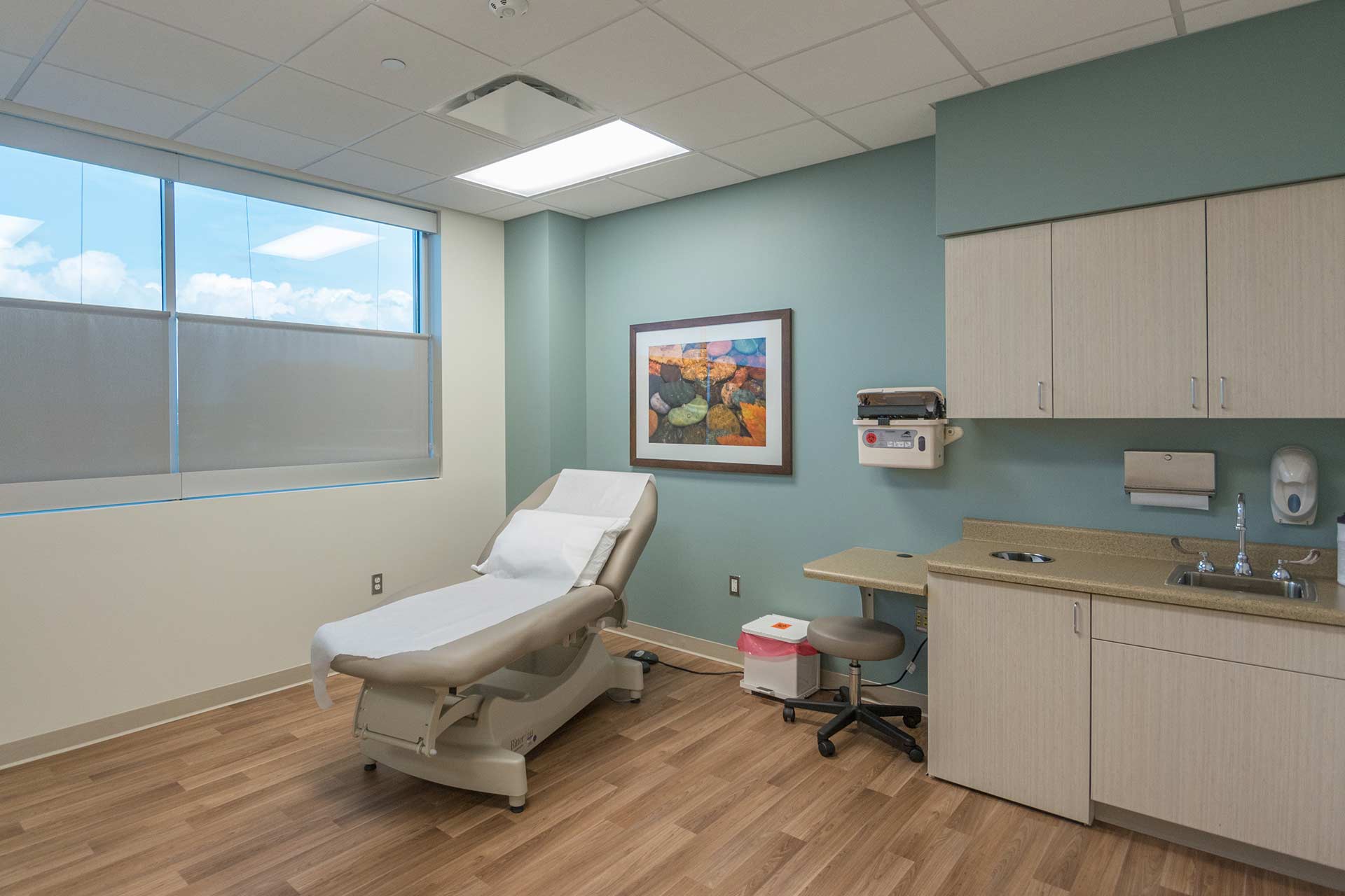 Modern exam room with patient chair, sink, cabinetry, and natural light from a large window.