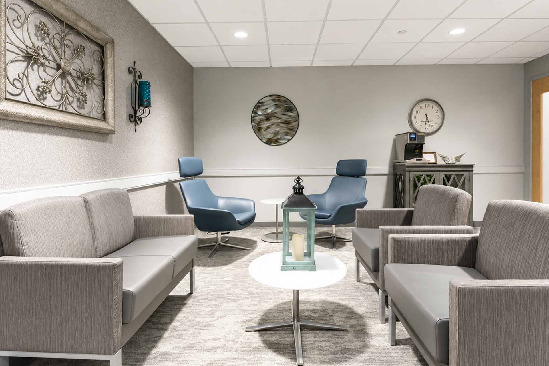 Comfortable medical waiting room with gray seating, blue accent chairs, coffee station, and modern décor.