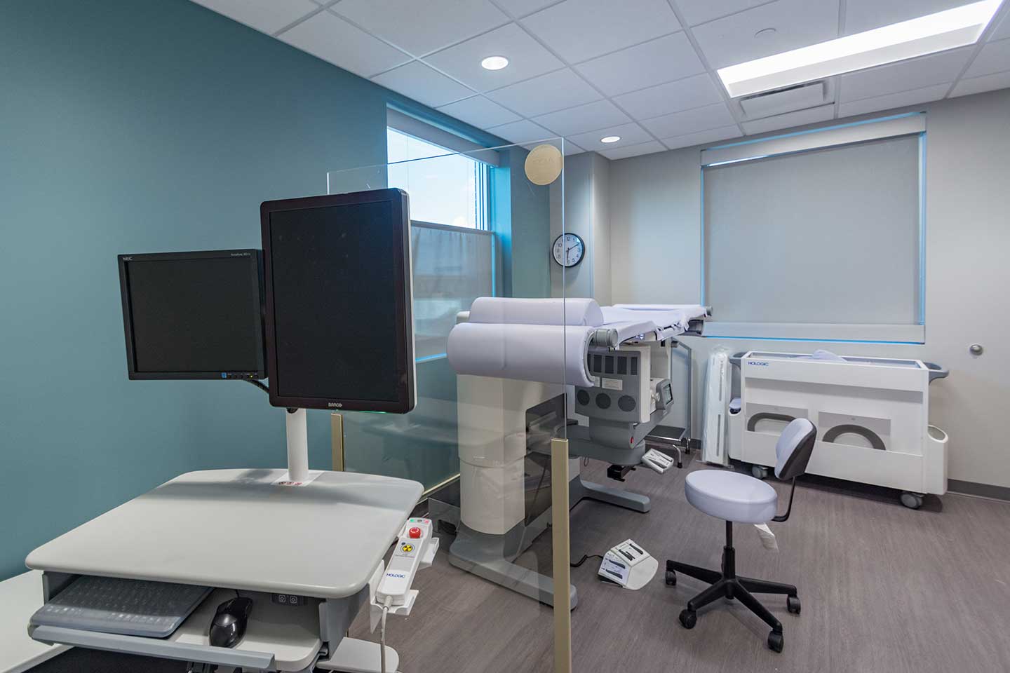 Medical imaging room with diagnostic equipment, dual monitors, exam chair, and patient examination table.