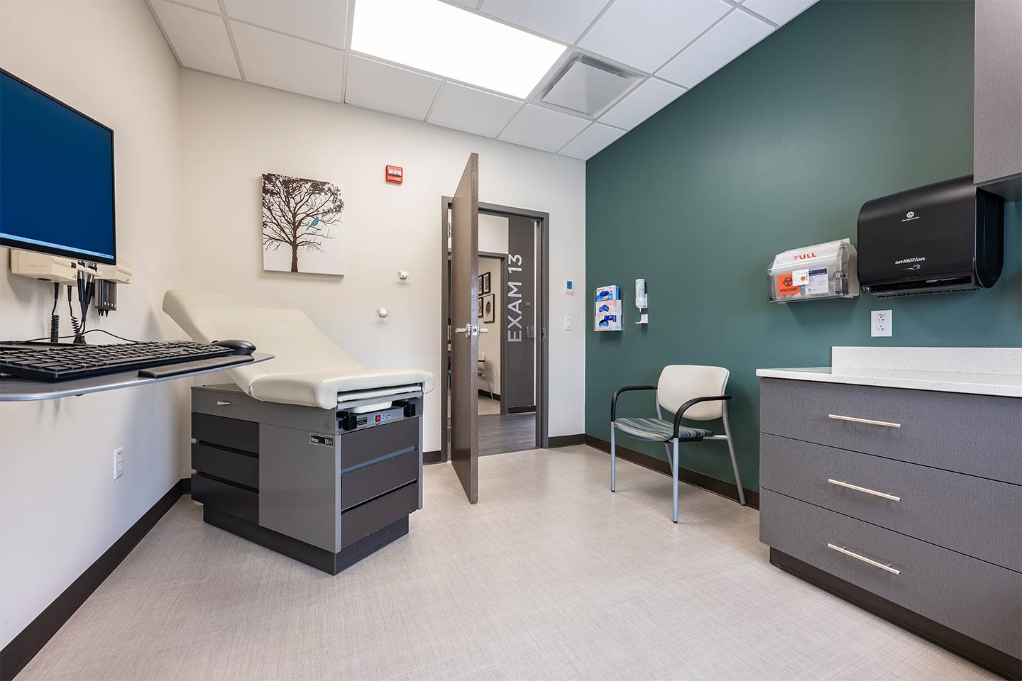 Modern medical exam room with exam table, computer workstation, patient chair, and storage cabinets.