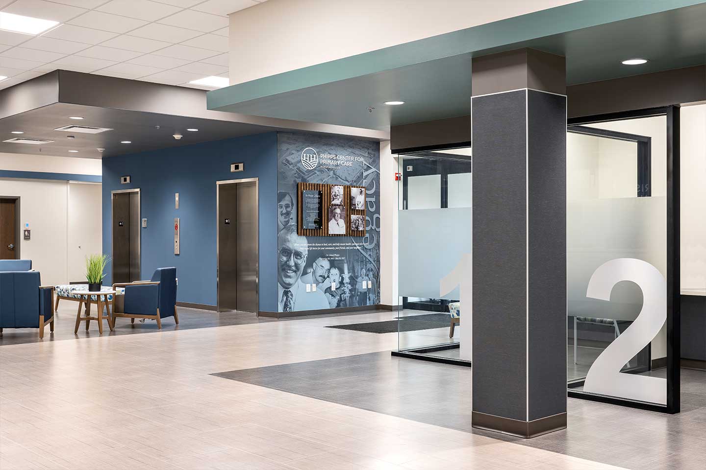 Modern healthcare lobby with elevators, seating area, glass meeting room, and history wall display.