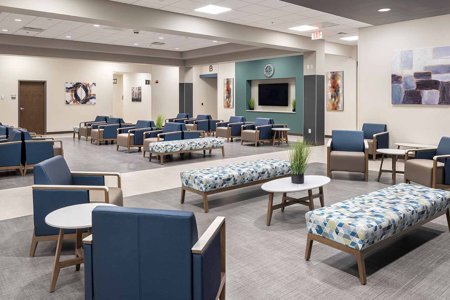 Spacious healthcare waiting area with comfortable seating, benches, tables, artwork, and a central TV display.