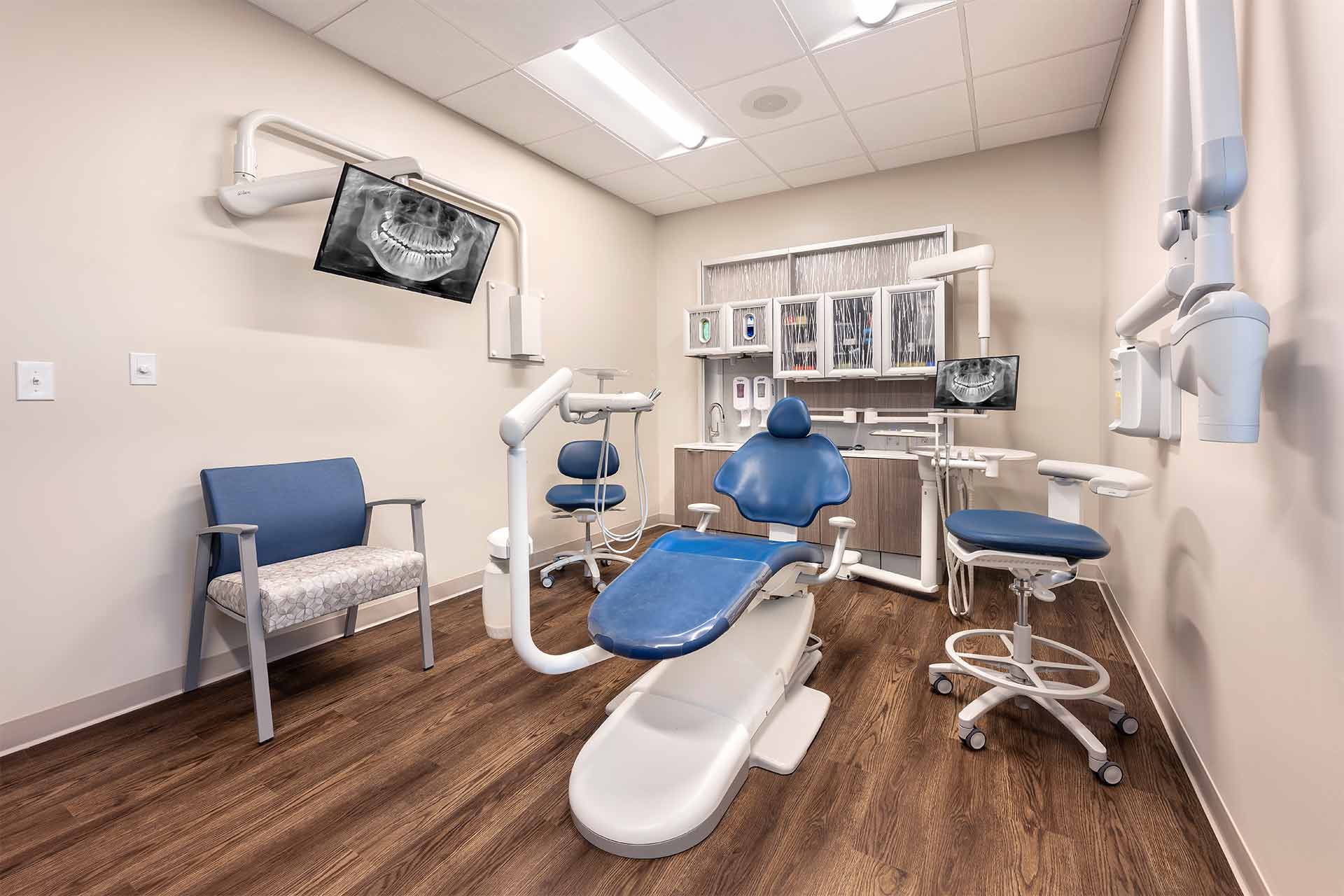 Dental exam room with blue dental chair, imaging monitors, and clinical equipment.