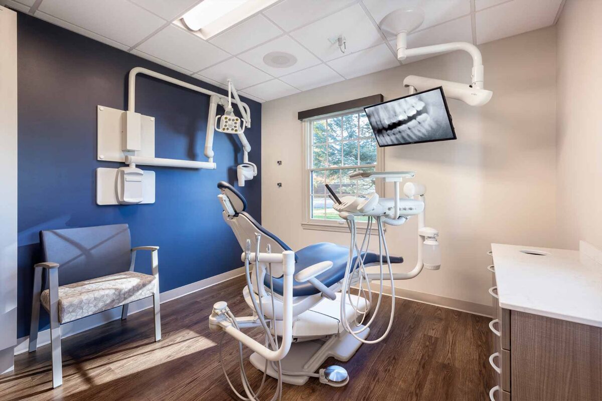 Modern dental exam room with treatment chair, overhead light, X-ray display, and large window.