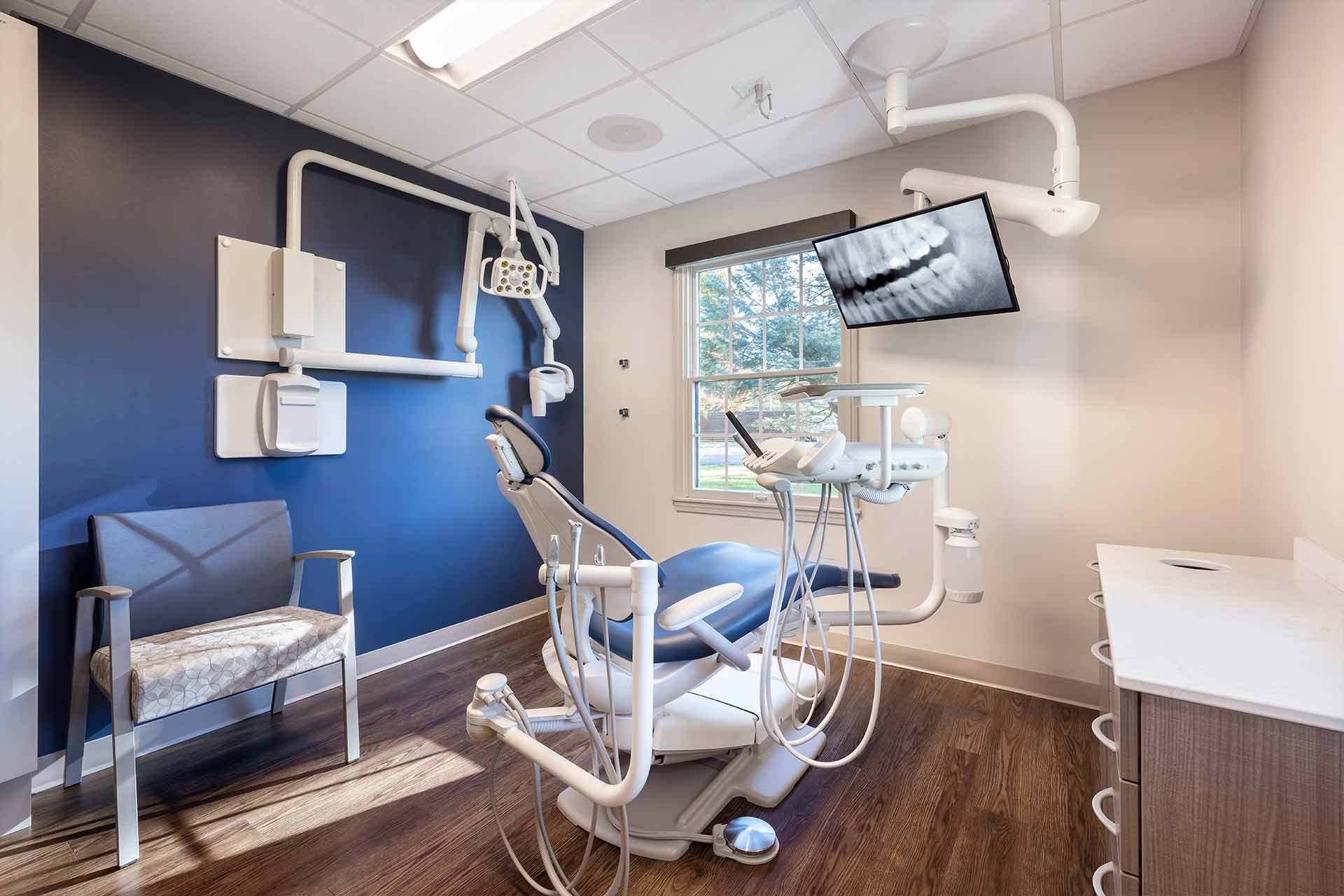 Modern dental exam room with treatment chair, overhead light, X-ray display, and large window.