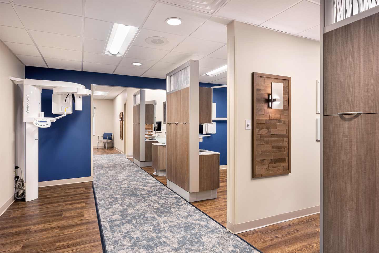 Modern dental office hallway with blue accent walls, wood finishes, treatment bays, and a panoramic X-ray machine.