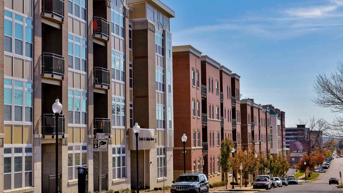 Street-level view of multi-story modern brick and glass apartment complex along a city road lined with parked cars.