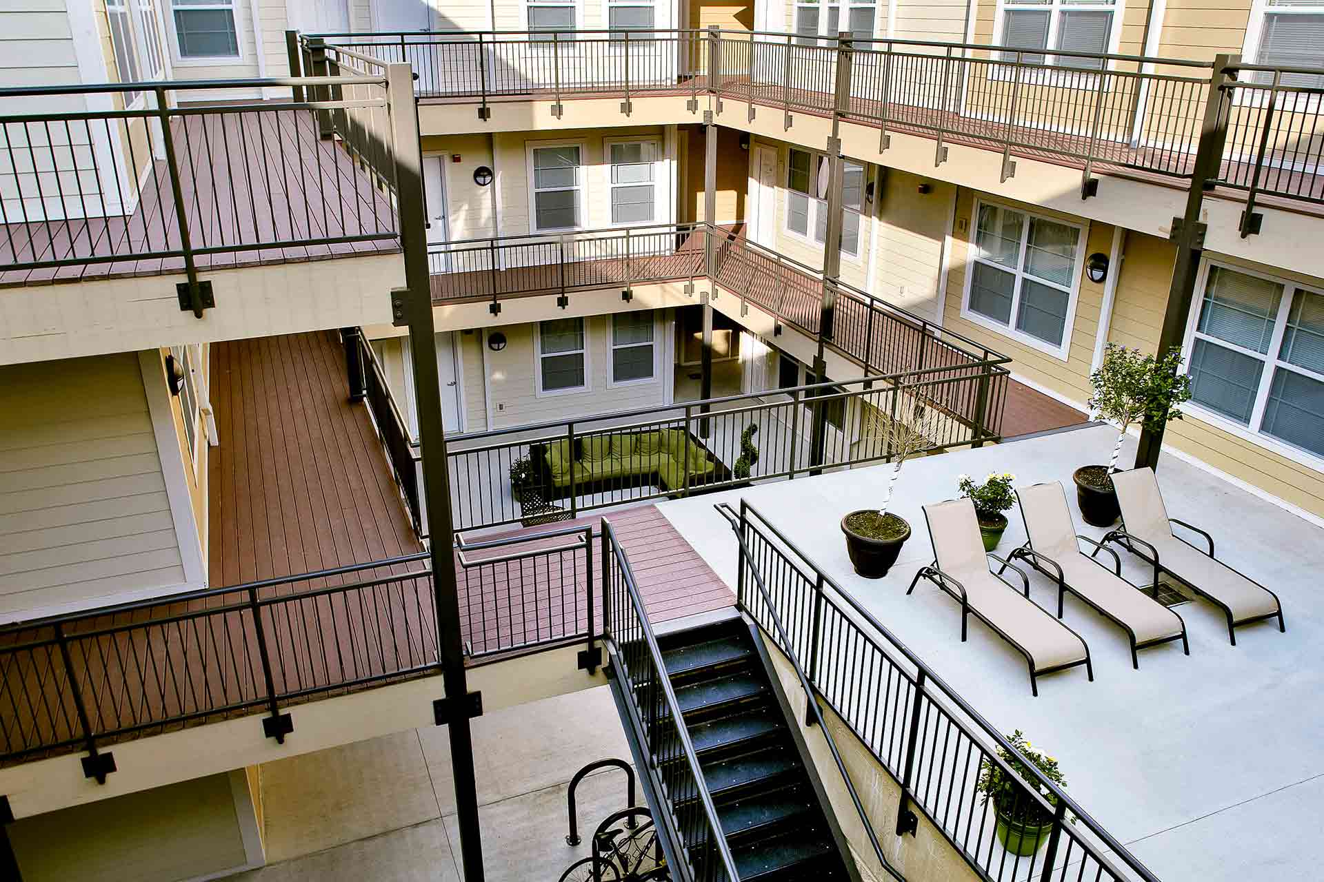 Multi-level outdoor apartment courtyard featuring walkways, railings, lounge chairs, and potted plants.