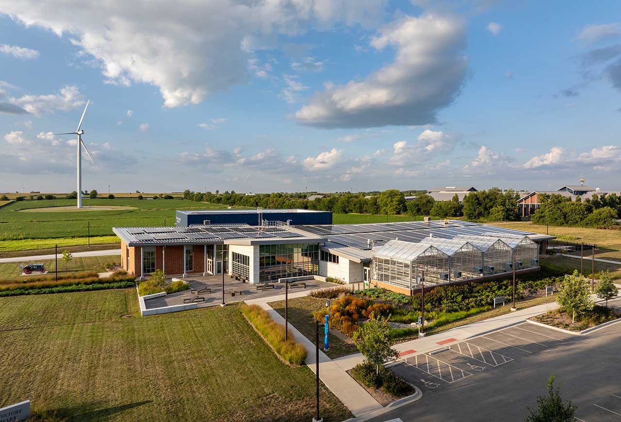 Legat-designed Heartland Community College Net Zero Agriculture Complex featuring solar panels, greenhouse, farmland, and a nearby wind turbine.